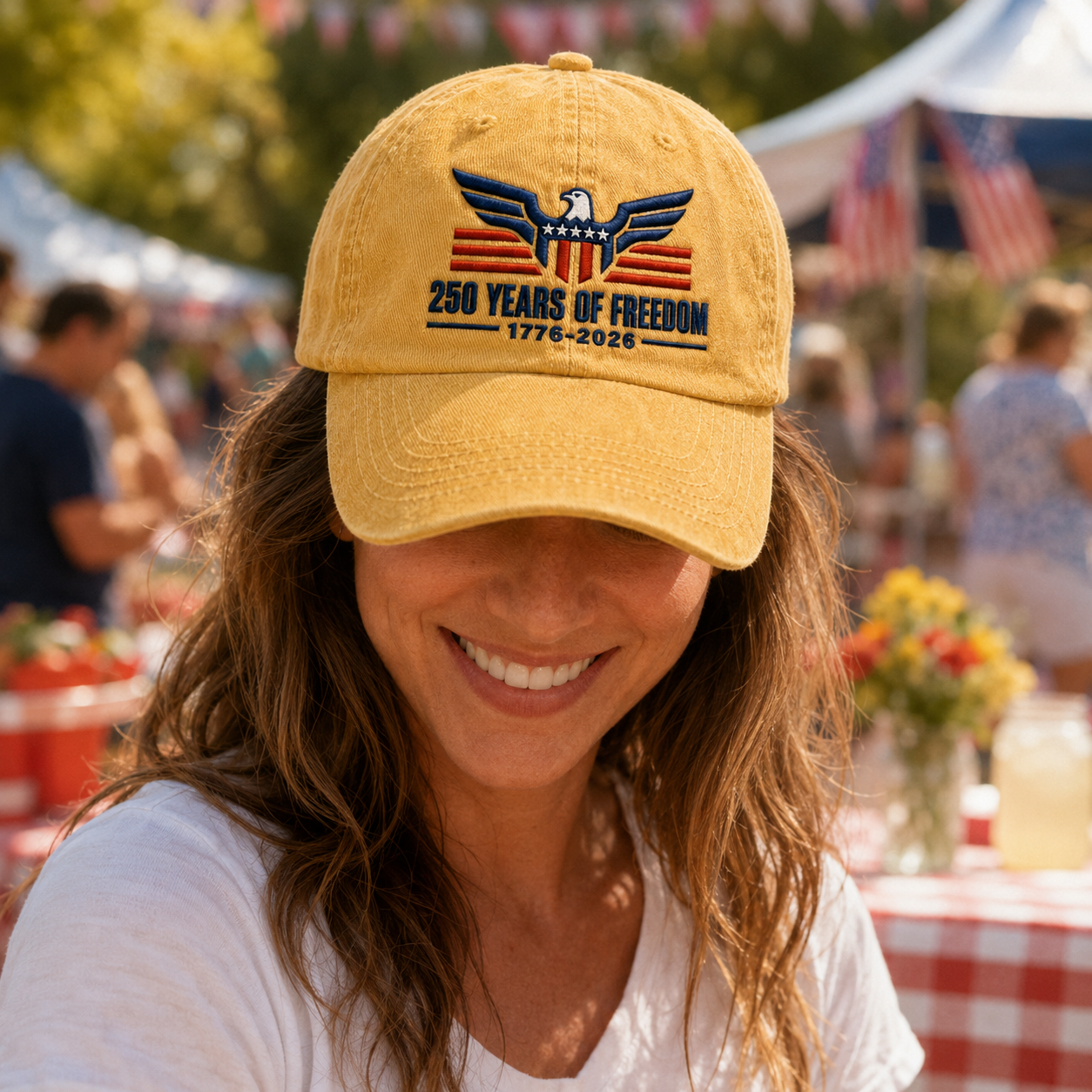 Woman at an outdoor gathering wearing a yellow washed baseball cap with embroidered eagle and 1776-2026 text