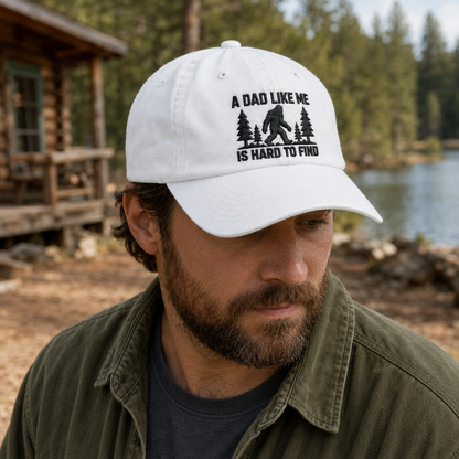 Man next to a lake cabin wearing a white washed cotton baseball cap with black embroidered bigfoot artwork