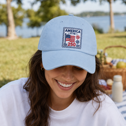 Woman smiling at a picnic wearing a light blue washed baseball cap with an embroidered patriotic America 250 stamp design