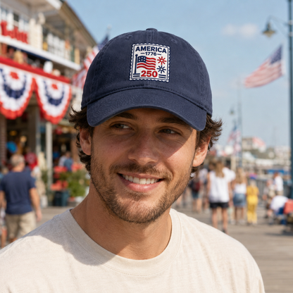 Man smiling on a boardwalk wearing a navy washed baseball cap with an embroidered patriotic America 250 stamp design