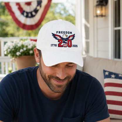 Man looking down while sitting on a porch wearing a white washed baseball cap with embroidered patriotic freedom 250 eagle design