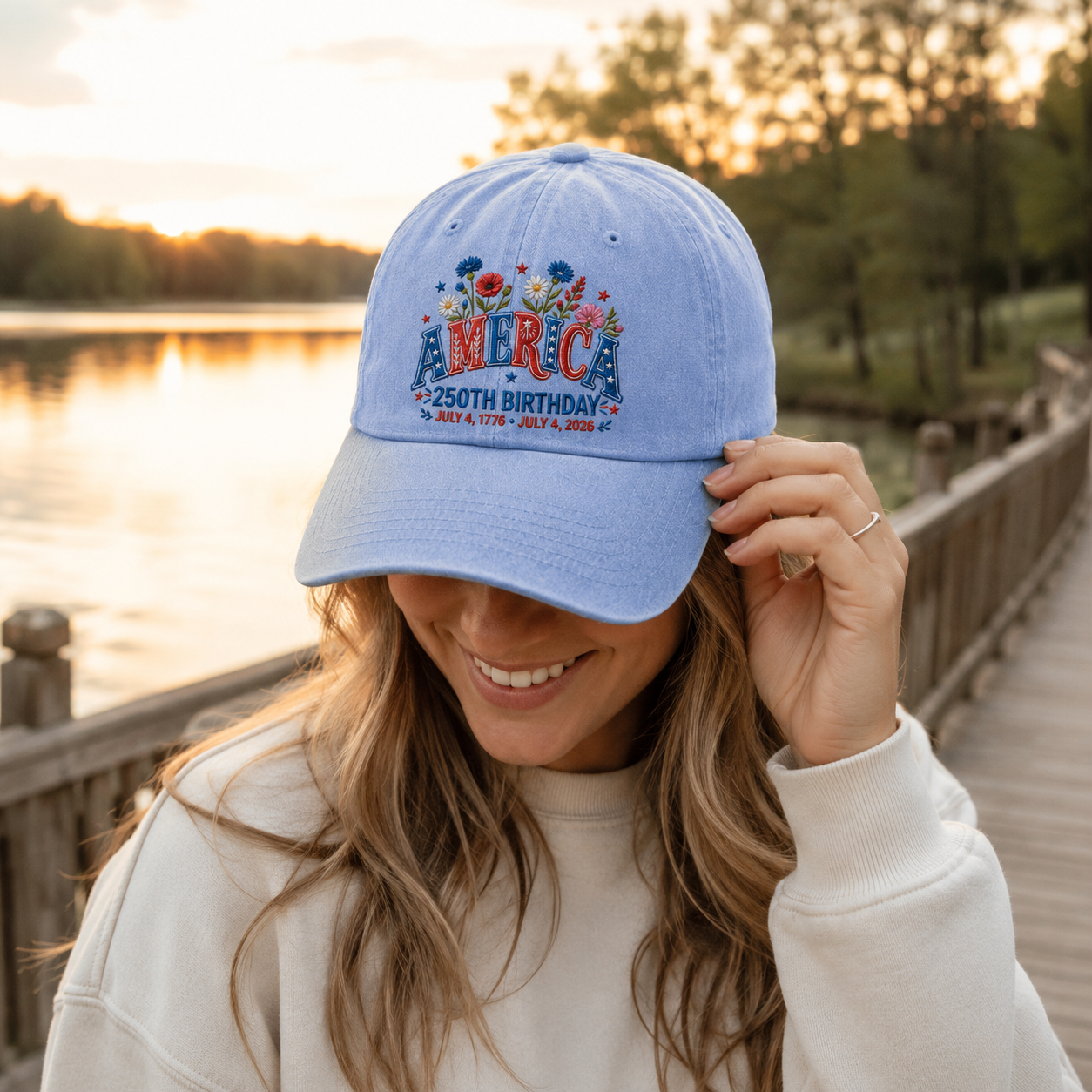 Woman adjusting a light blue washed baseball cap with patriotic floral America embroidery by a lake at sunset