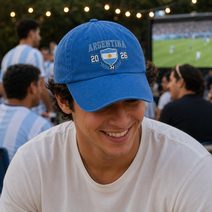 Man smiling at a tournament watch party with a big screen wearing a royal blue washed baseball cap with an embroidered Argentina 2026 soccer crest design