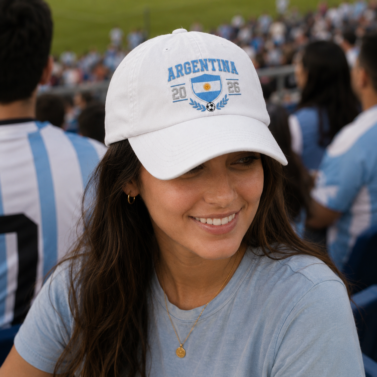 Woman smiling in a stadium grandstand wearing a white washed baseball cap with an embroidered Argentina 2026 soccer crest design