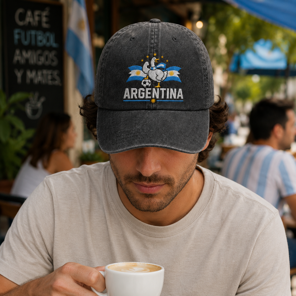 Man drinking coffee at an outdoor café wearing a black washed baseball cap with an embroidered Argentina soccer goose design