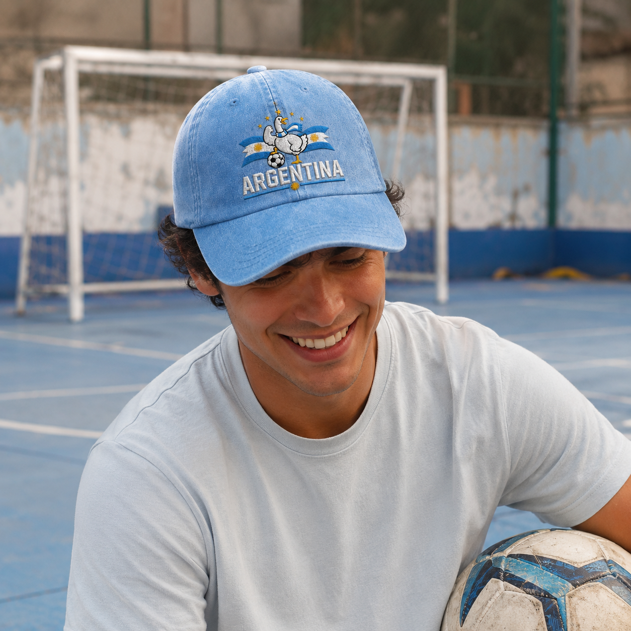 Man holding a football on a blue sports court wearing a light blue washed baseball cap with an embroidered Argentina soccer goose design