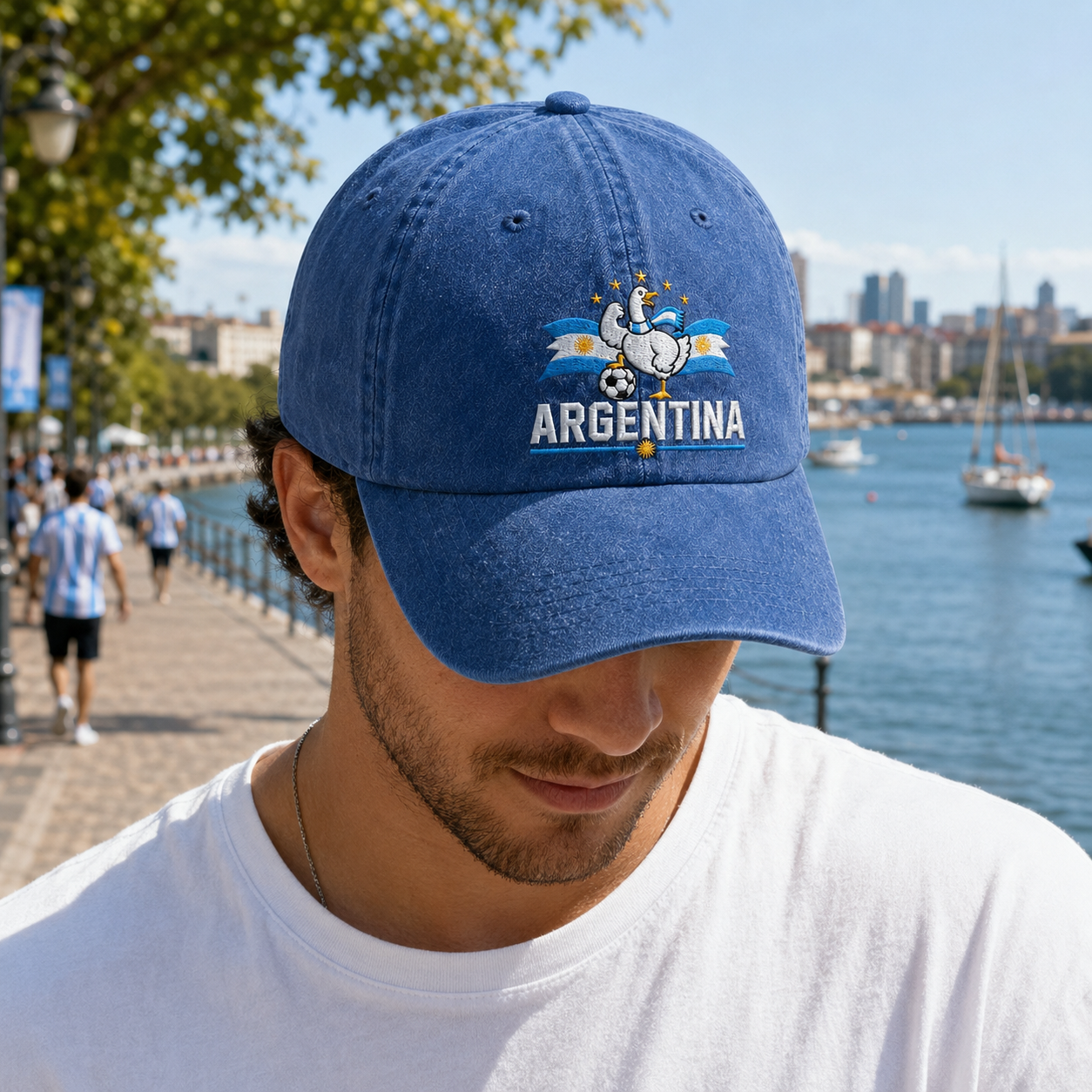 Man looking down on a waterfront boardwalk wearing a blue washed baseball cap with an embroidered Argentina soccer goose design