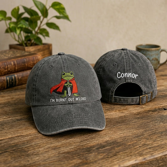 Two gray baseball caps on a wooden surface with one featuring a frog graphic and text, the other with 'Connor' embroidered.