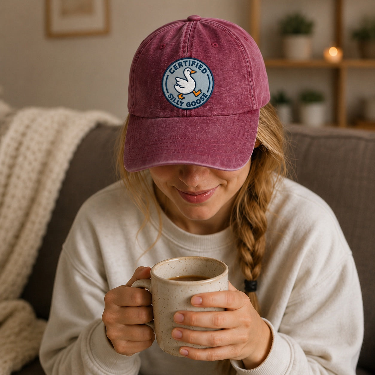 Person wearing a maroon cap with a logo, holding a mug in a cozy indoor setting.