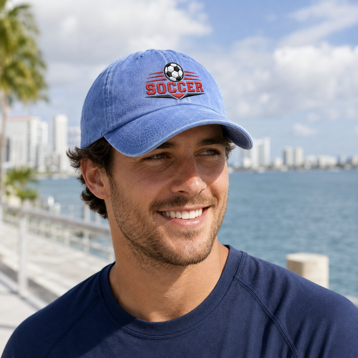 Man smiling on a coastal city waterfront walkway wearing a light blue washed baseball cap with an embroidered graphic soccer ball logo and red text
