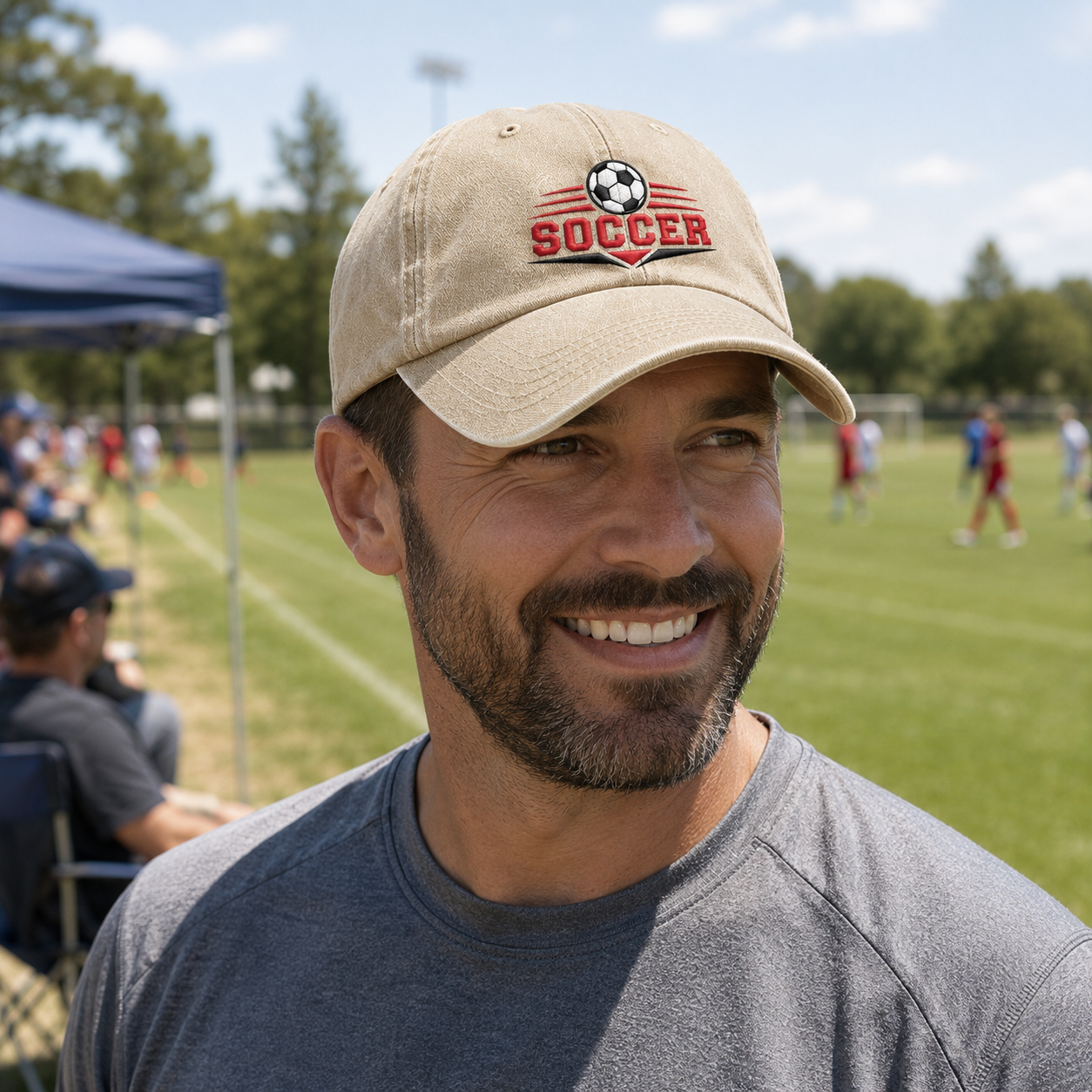 Man smiling by a sports field sideline dugout wearing a sand washed baseball cap with an embroidered graphic soccer ball logo and red text