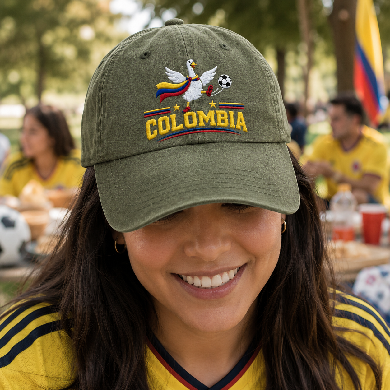 Woman smiling at an outdoor game day watch party wearing an army green washed baseball cap with an embroidered Colombia soccer goose design