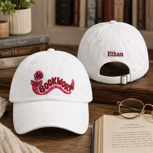 Front and back view of a white washed cotton baseball cap next to an open book featuring an embroidered pink cartoon bookworm design and custom name Ethan text