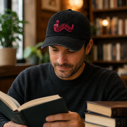 Man reading a book in a library setting wearing a black washed baseball cap with an embroidered pink cartoon bookworm design