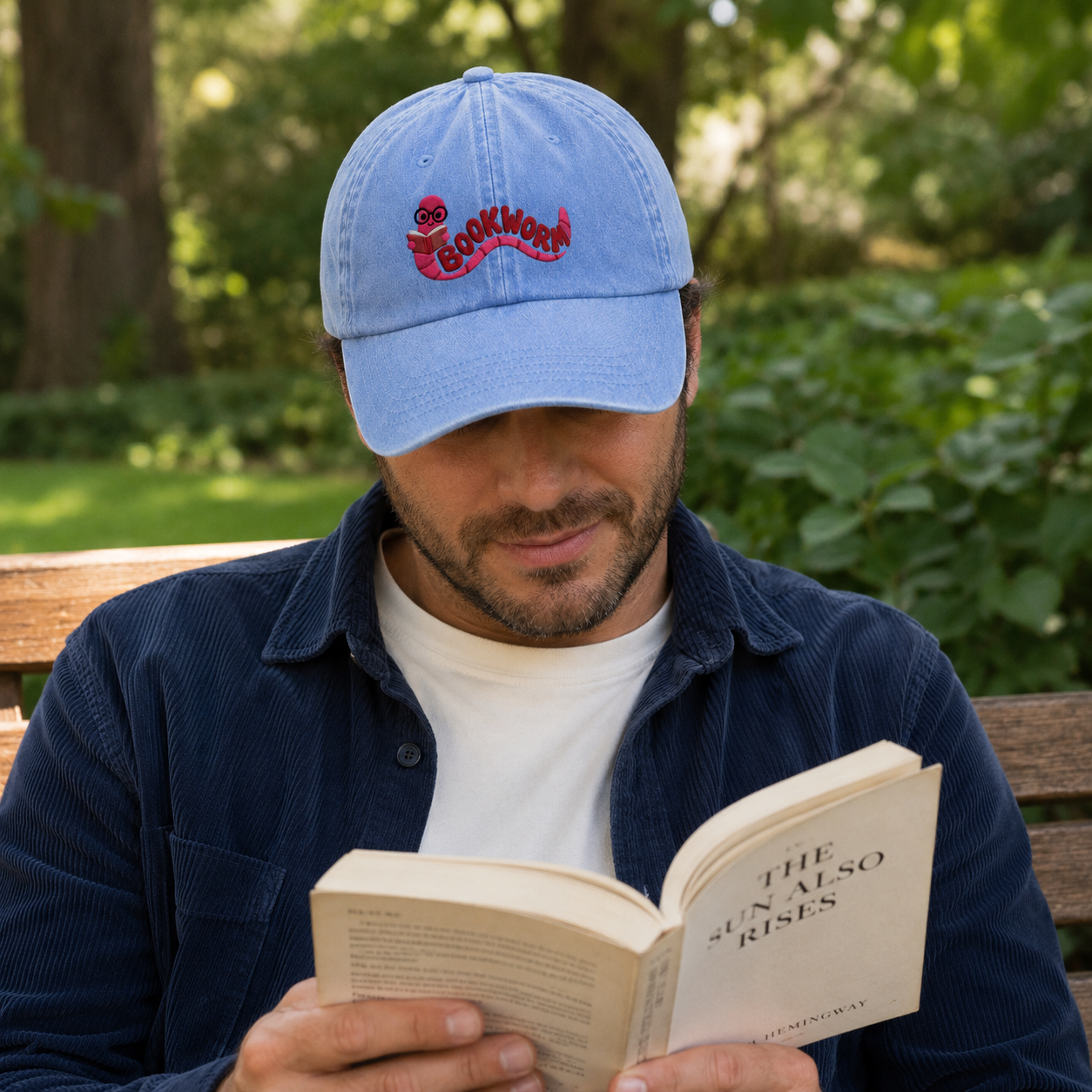 Man reading on a park bench wearing a light blue washed baseball cap with an embroidered pink cartoon bookworm design