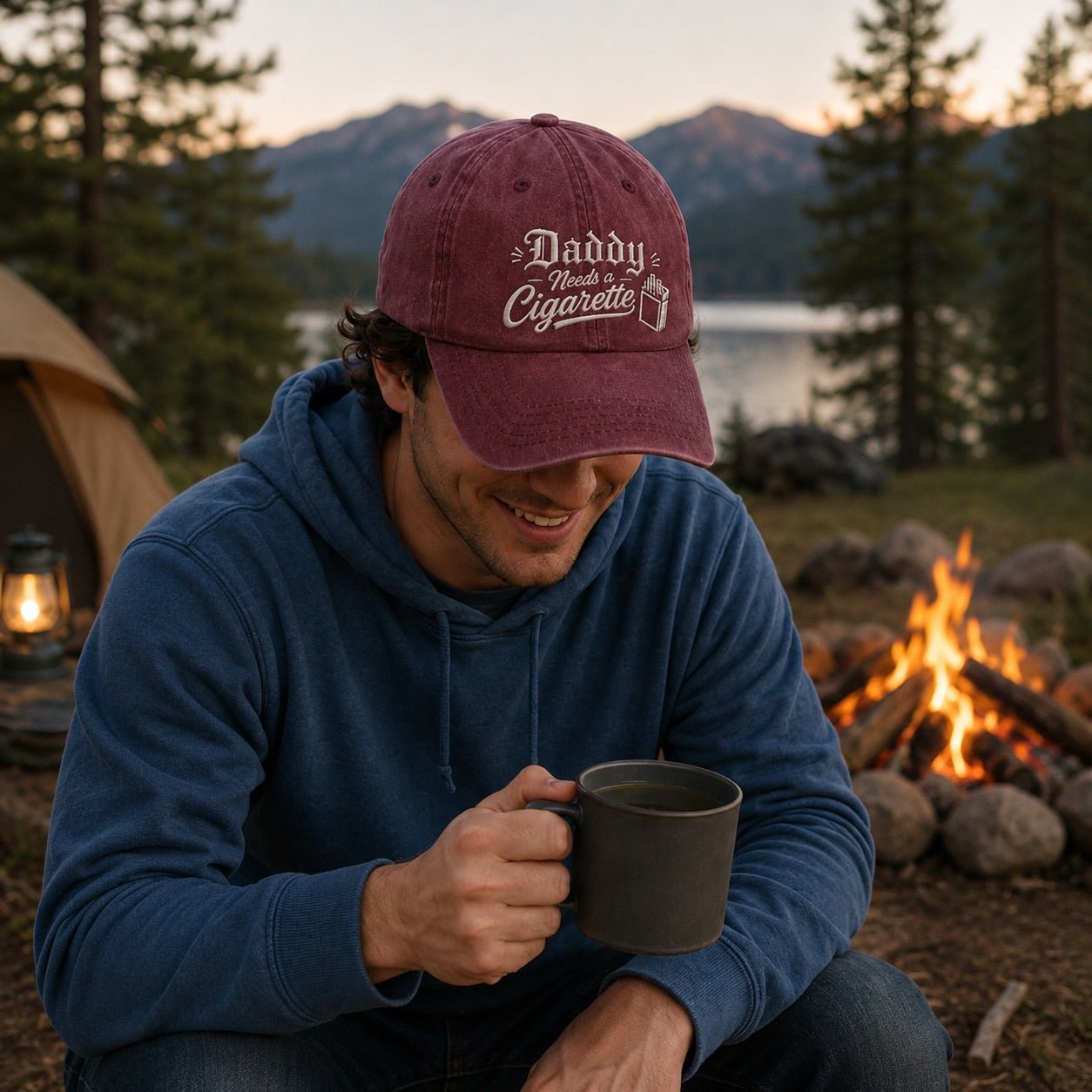 Man wearing a 'Daddy Needs Cigarettes' cap by a campfire with mountains in the background