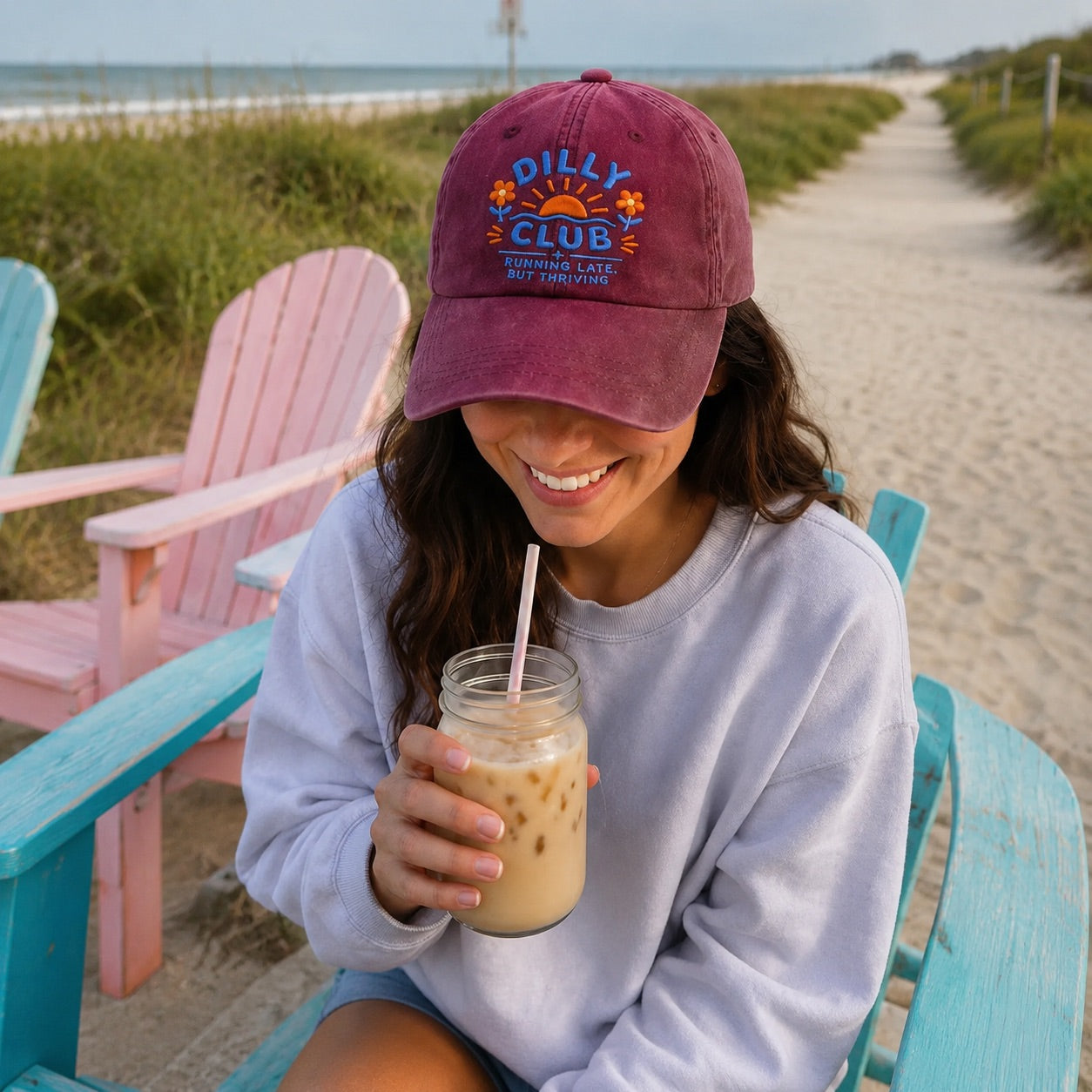 Woman sitting on a beach with a 'Dilly Club' cap and iced coffee.