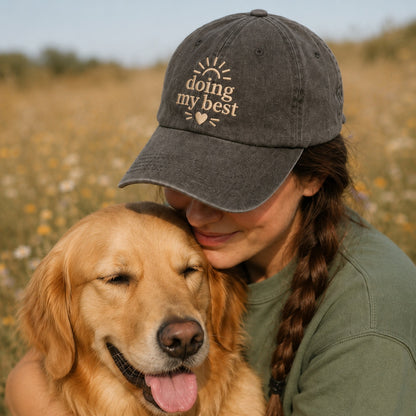 Woman wearing a cap with 'doing my best' text, hugging a golden dog in a field.