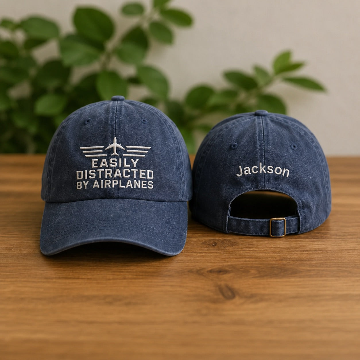 Two navy blue baseball caps with text on a wooden surface with a plant in the background