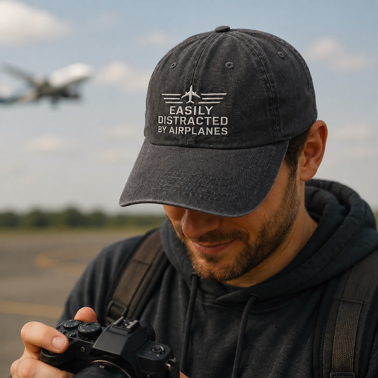 Man wearing a cap with 'Easily Distracted by Airplanes' text, holding a camera on an airfield.