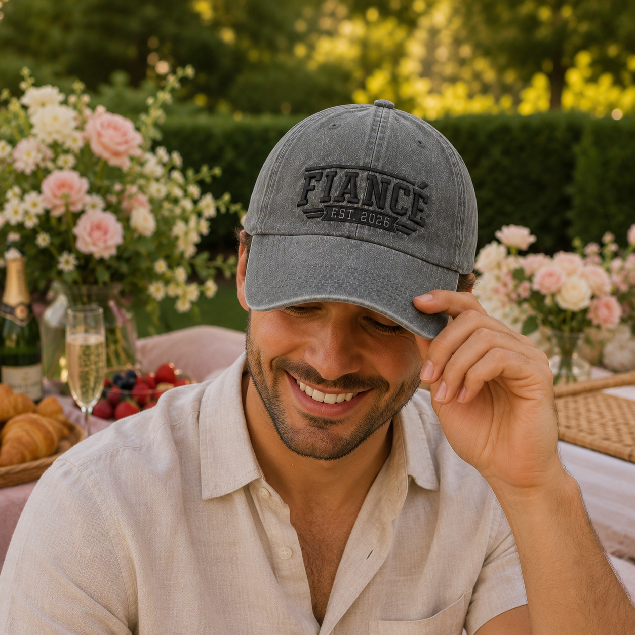 Smiling man at a garden picnic table wearing a grey washed baseball cap with embroidered black Fiance Est 2026 text
