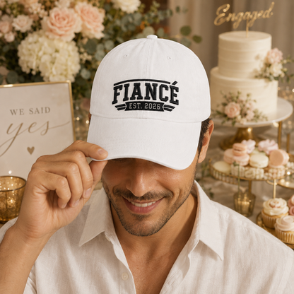Smiling man next to a celebratory engagement cake wearing a white washed baseball cap with embroidered black Fiance Est 2026 text