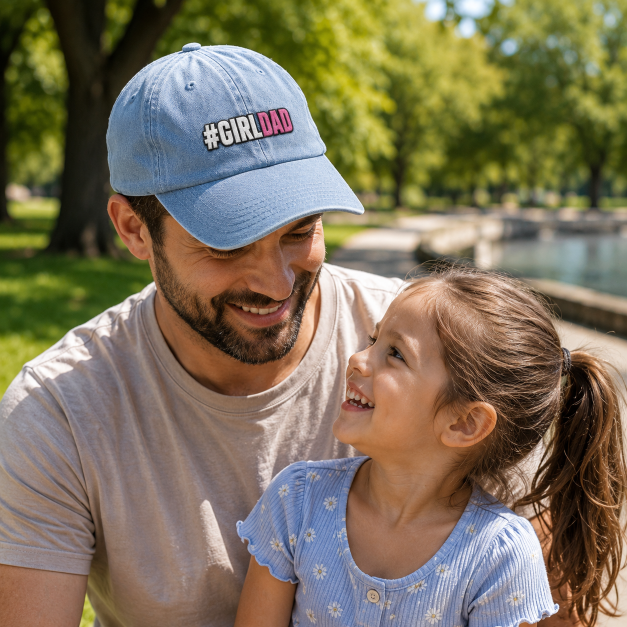 Father and daughter smiling at each other in a park while he wears a light blue washed embroidered baseball cap