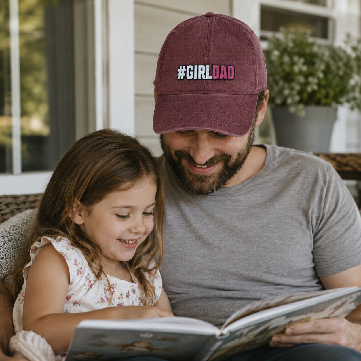 Father and daughter reading a book together while he wears a maroon washed baseball cap with embroidered girl dad text