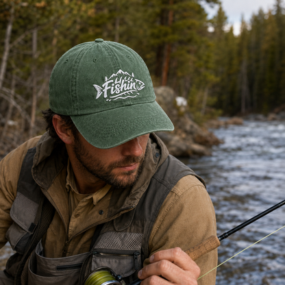 Man fly fishing by a rushing mountain river wearing an army green washed baseball cap with white embroidered Fishin mountain design