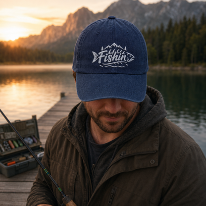 Man holding a fishing rod on a wooden pier at sunset wearing a navy washed baseball cap with white embroidered Fishin mountain design