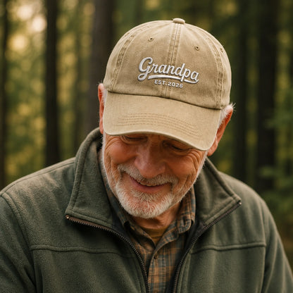 Man wearing a beige cap with 'Grandpa Est. 2026' text, standing in a forest.
