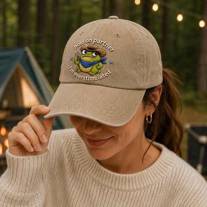 Person wearing a beige cap with a frog graphic and text, standing in a forest setting with tents.