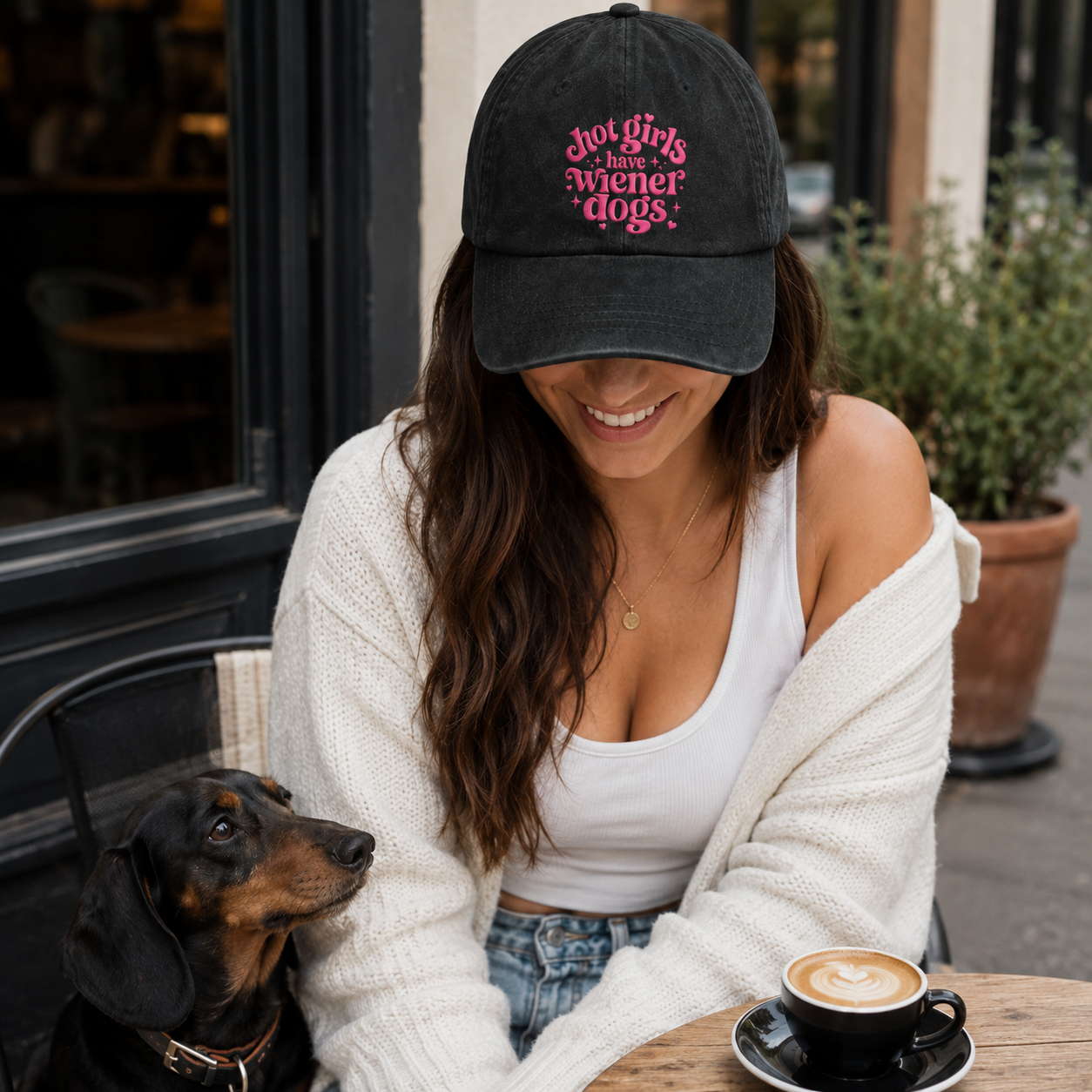 Woman at an outdoor cafe with a dachshund wearing a black washed baseball cap with pink retro text quote