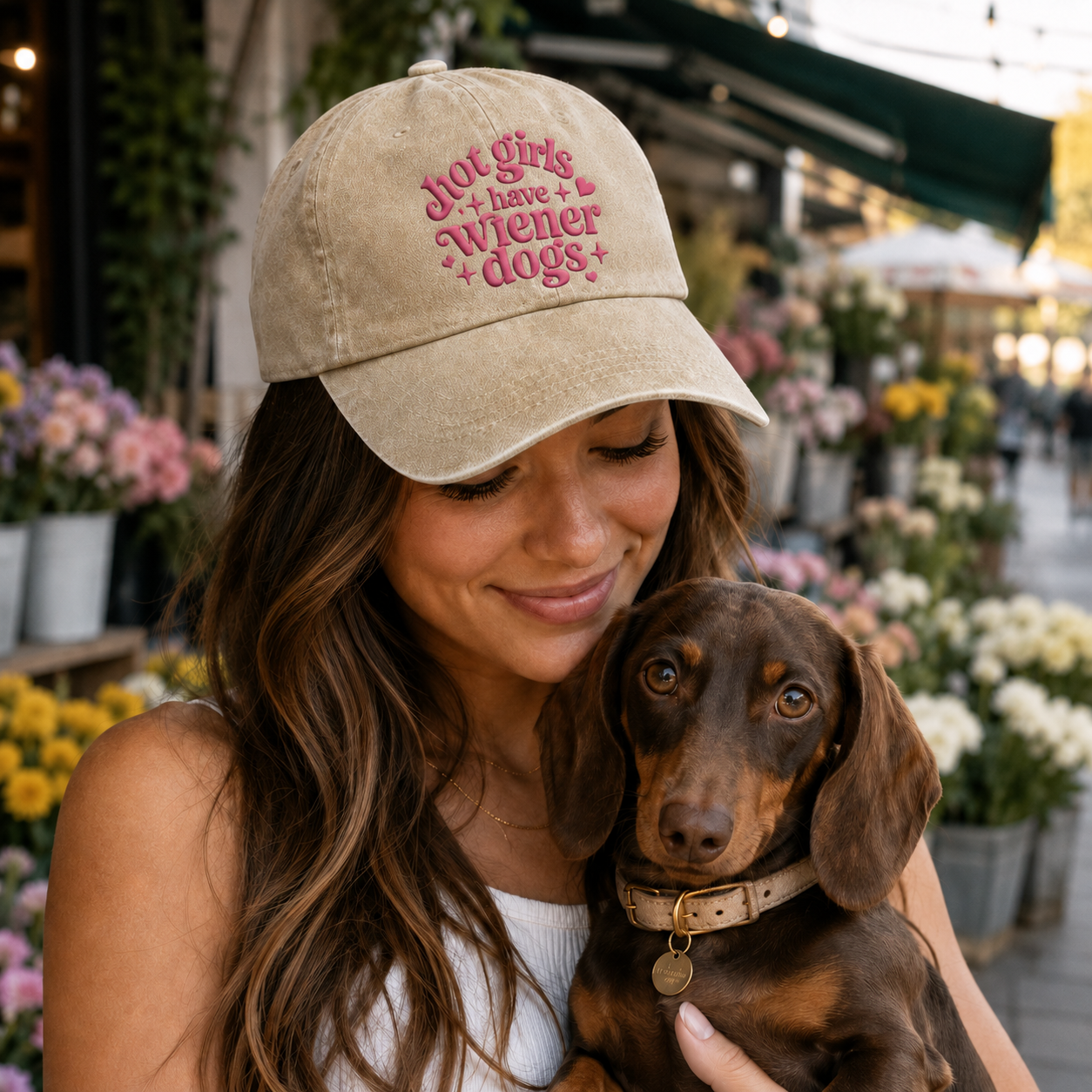 Woman holding a dachshund at a flower market wearing a sand washed baseball cap with pink retro text quote