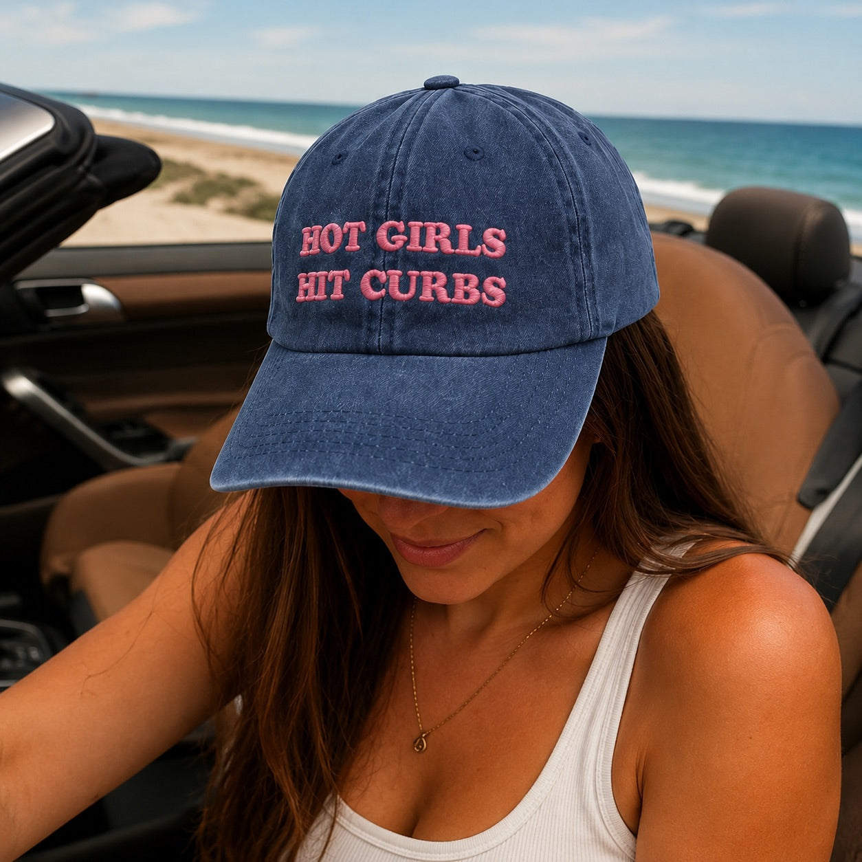 Person wearing a blue cap with 'HOT GIRLS HIT CURBS' text in a car by the beach.
