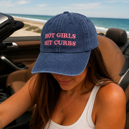 Person wearing a blue cap with 'HOT GIRLS HIT CURBS' text in a car by the beach.