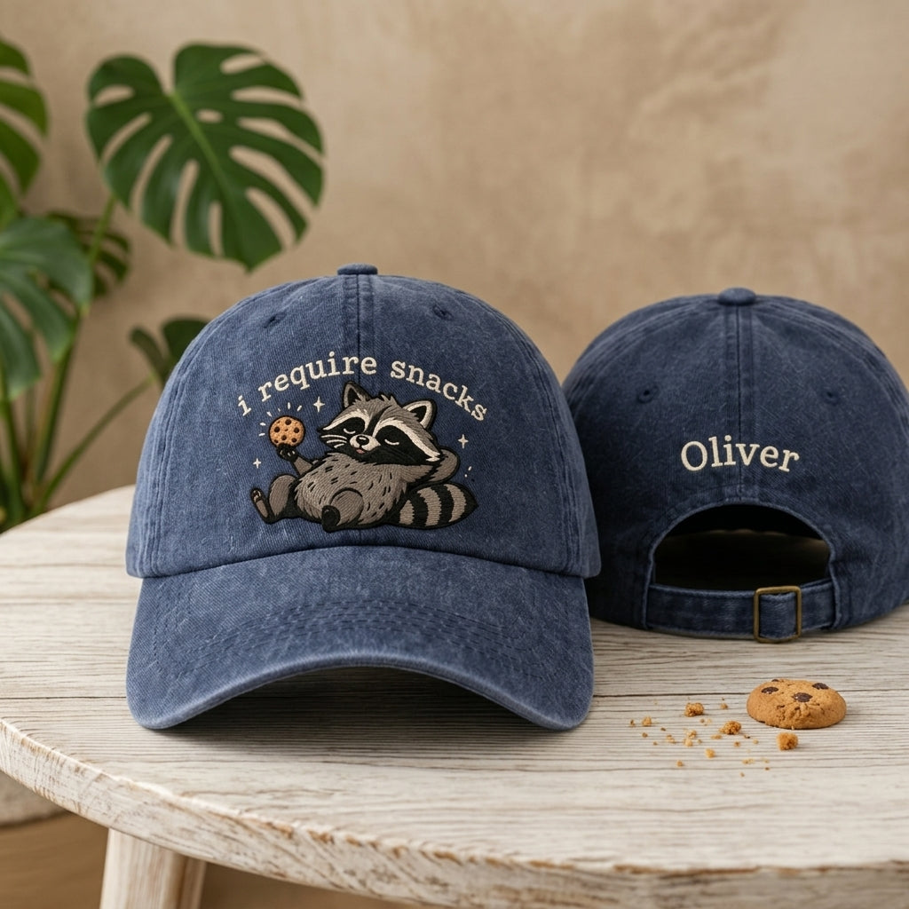Blue baseball cap with raccoon design and 'i require snacks' text on a wooden table.
