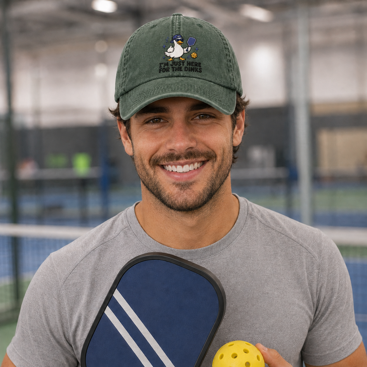 Man smiling indoors holding a paddle and a yellow ball, wearing an army green washed baseball cap with an embroidered cartoon duck pickleball design