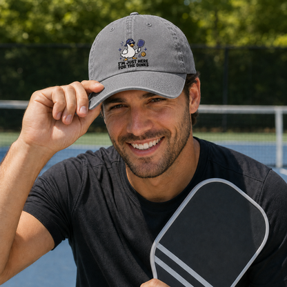 Man adjusting his cap on a court while holding a paddle, wearing a grey washed baseball cap with an embroidered cartoon duck pickleball design