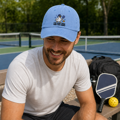 Man smiling on a court bench next to a paddle and backpack, wearing a light blue washed baseball cap with an embroidered cartoon duck pickleball design