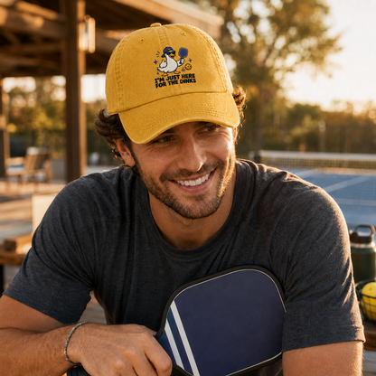Man smiling on an outdoor court while holding a paddle, wearing a yellow washed baseball cap with an embroidered cartoon duck pickleball design