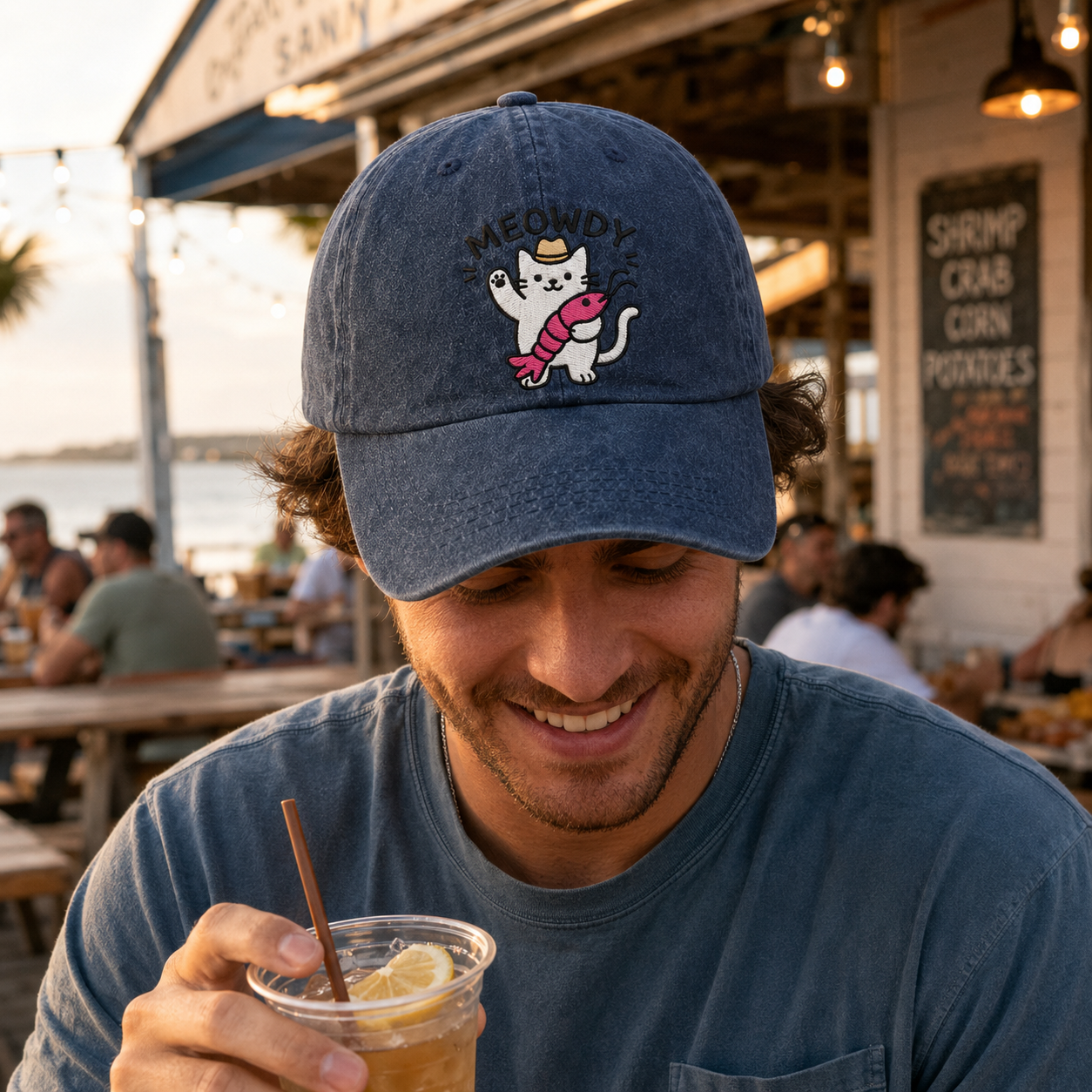 Smiling man drinking a beverage at a coastal restaurant wearing a blue washed baseball cap with an embroidered cartoon cat holding a crawfish