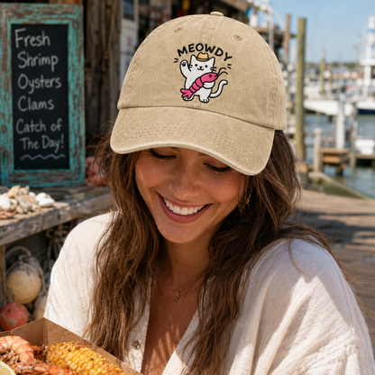 Smiling woman holding a seafood boil platter at a pier wearing a sand washed baseball cap with an embroidered cartoon cat holding a crawfish