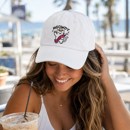 Smiling woman holding a cold beverage outdoors wearing a white washed baseball cap with an embroidered cartoon cat holding a crawfish