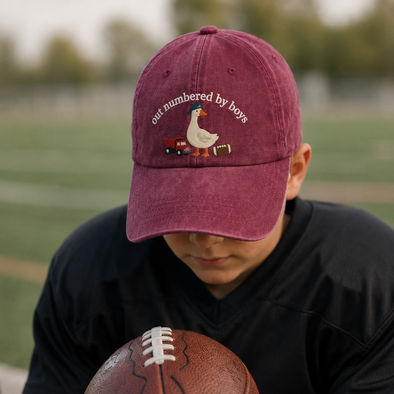Person wearing a maroon cap with a duck graphic and text, holding a football on a grassy field.