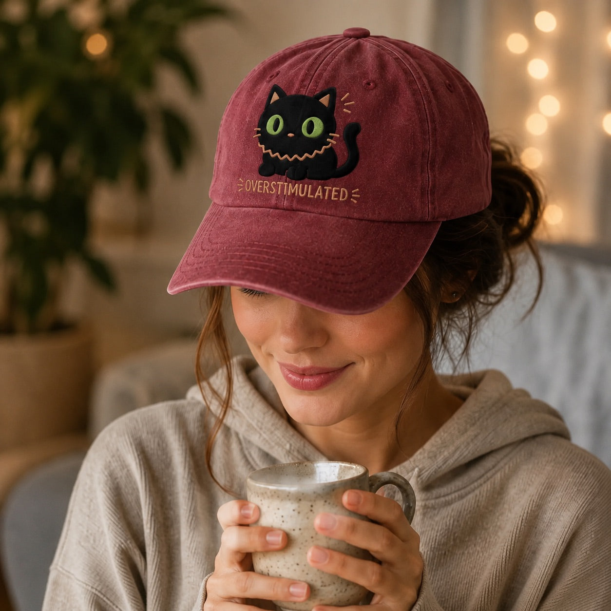 Person wearing a maroon cap with a cat design, holding a mug indoors.