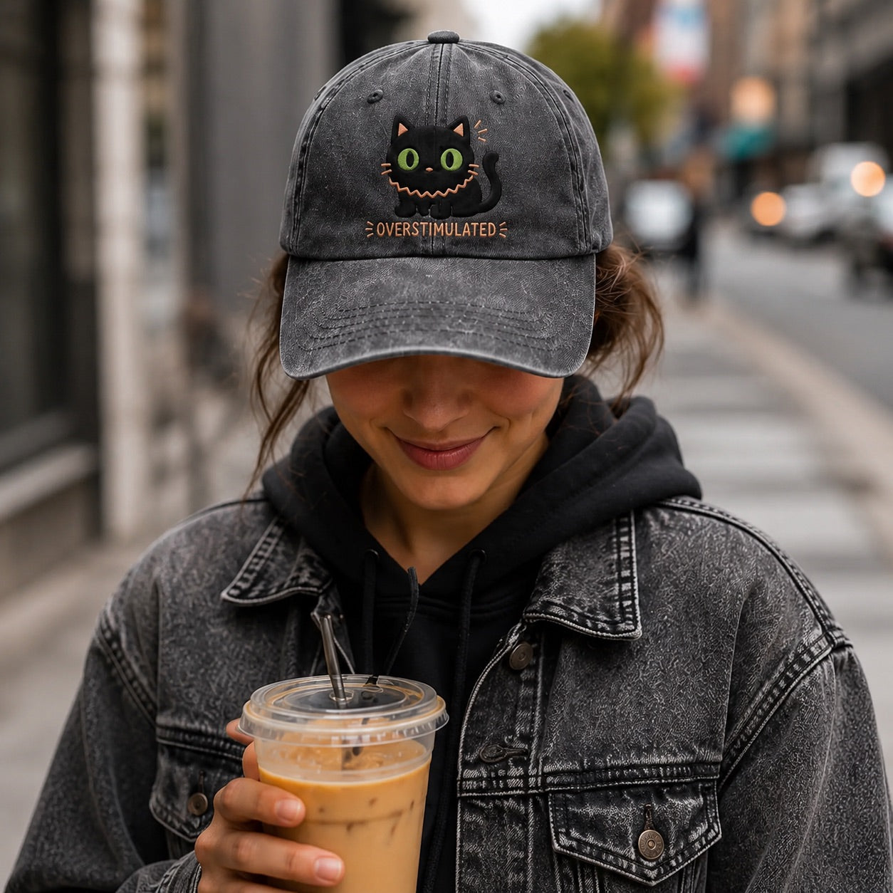 Person wearing a cap with a cat design and holding a drink on a city street.