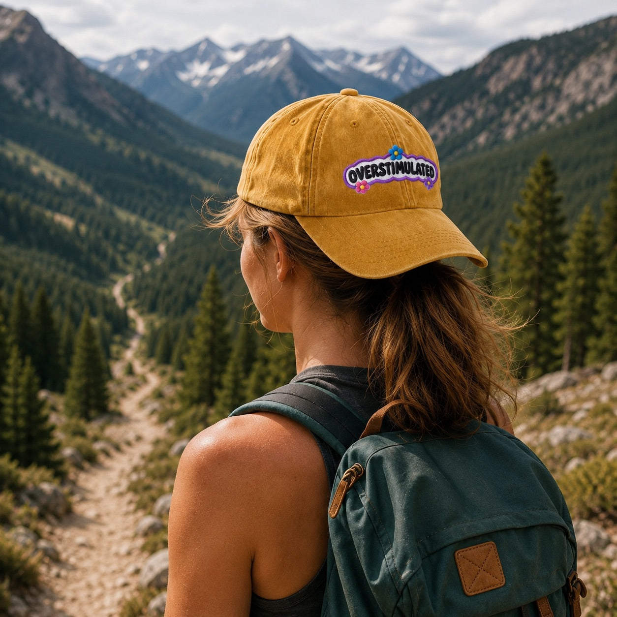 Person wearing a yellow cap with 'Overstimulated' logo and green backpack, standing in a mountainous landscape.