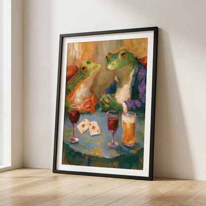 A framed art print leaning against a wall on a light wood floor. The print shows two green frogs seated at a table with drinks and playing cards, rendered in a painterly, colorful style.