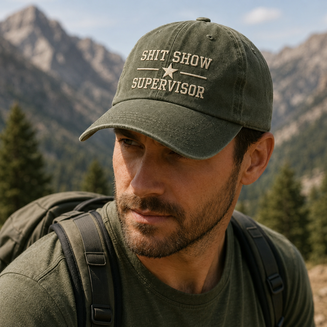 Man wearing a cap with 'Shit Show Supervisor' text in a mountainous landscape